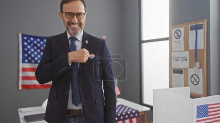 Hombre barbudo de mediana edad mostrando orgullosamente la etiqueta 'voté' en el entorno de la universidad electoral estadounidense con banderas