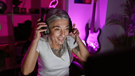 Foto de Confiado jugador femenino de mediana edad, streamer de pelo gris con auriculares en la sala de juegos oscura, sonriendo mientras transmite videojuegos en línea - Imagen libre de derechos