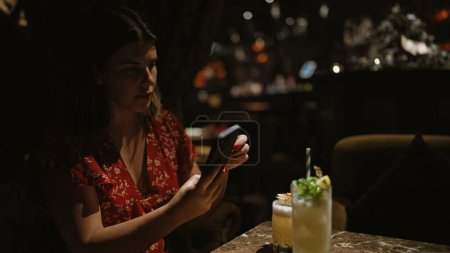 Beautiful hispanic woman captures fun nightlife moment, joyously tableside holding tropical cocktail drink at nightclub summer celebration