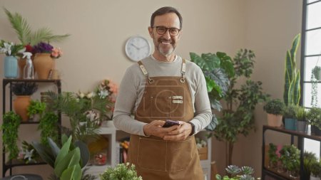 Foto de Hombre mayor sonriente con barba usando delantal usando teléfono inteligente en la vibrante tienda de flores - Imagen libre de derechos
