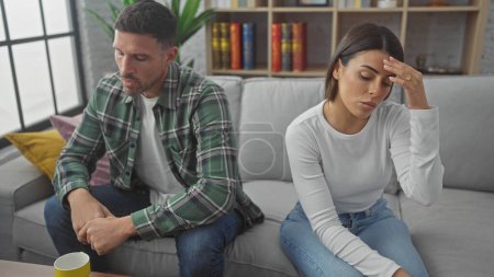 Ein besorgtes hispanisches Paar sitzt getrennt auf dem Sofa in einem modernen Wohnzimmer und spiegelt Spannung und Besorgnis wider.