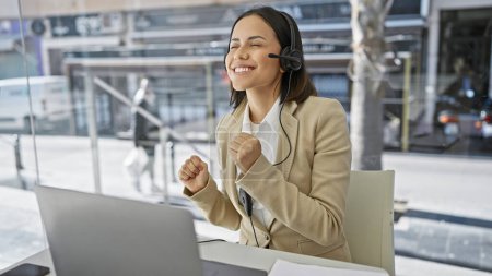 Photo for A cheerful young woman with a headset celebrates while using a laptop in a modern office with a blurred urban background. - Royalty Free Image