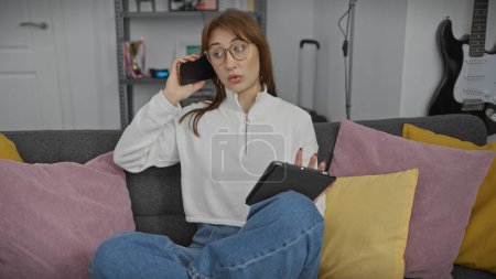 Photo for Caucasian woman talks on phone and holds tablet while sitting on sofa in a modern living room - Royalty Free Image