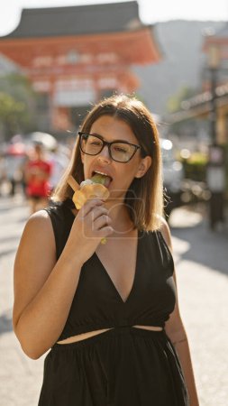 Cheerful hispanic woman savors deliciously sweet ice cream on kyoto street, her beautiful smile is framed by glasses while experiencing the traditional architecture of this japanese city