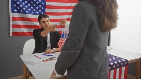 American woman candidate reaches to receive a ballot in a college electoral setting with the us flag present.