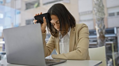 Foto de Una joven hispana estresada en un escritorio con un portátil sosteniendo su teléfono en su templo en un ambiente de oficina beige. - Imagen libre de derechos