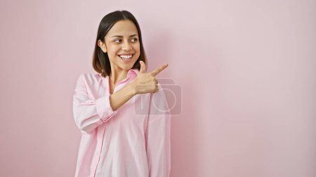 Foto de Mujer joven hispana sonriente con una camisa rosa apuntando hacia un lado sobre un fondo rosa liso. - Imagen libre de derechos