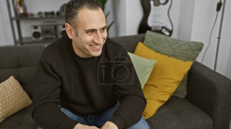 Hombre sonriente sentado cómodamente en un sofá gris en una acogedora sala de estar con almohadas de colores y un fondo de guitarra