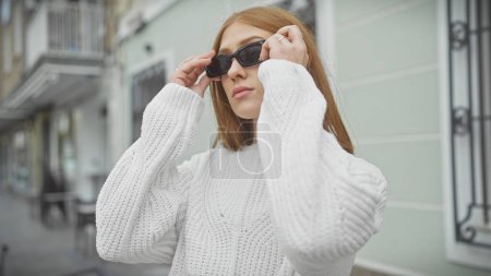 Foto de Caucásico pelirroja joven mujer ajustando gafas de sol en una calle de la ciudad, retratando el estilo urbano. - Imagen libre de derechos