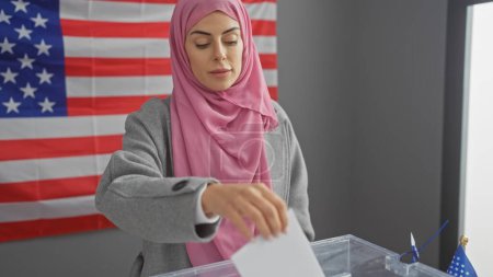 Una joven con un hiyab arroja una boleta electoral en un colegio electoral con una bandera americana en el fondo.