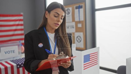 Mujer hispana vestida con calcomanía de 'yo voté' toma notas en un centro electoral con banderas americanas