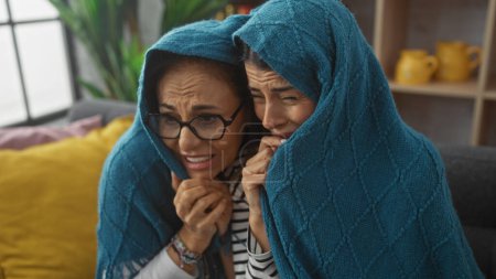 Foto de Dos mujeres compartiendo un momento emocional bajo una manta azul en un sofá en casa - Imagen libre de derechos