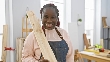 Foto de Mujer africana sonriente con trenzas usando un delantal en un estudio de carpintería brillante sosteniendo madera. - Imagen libre de derechos