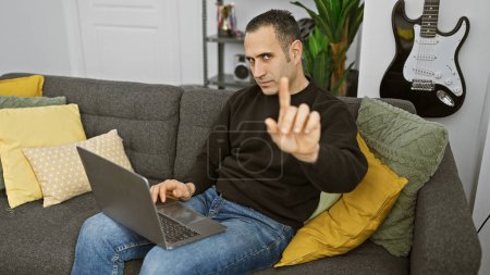Foto de Hombre hispano dando señal de desaprobación mientras trabaja en un ordenador portátil en una sala de estar moderna. - Imagen libre de derechos