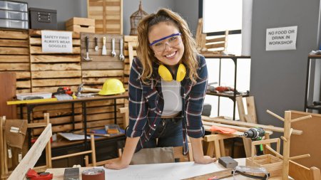 Foto de Sonriente joven hispana, una hermosa carpintera con gafas de seguridad brillando brillantemente en su acogedor taller de carpintería - Imagen libre de derechos