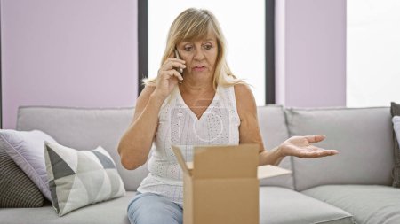 Upset middle-aged blonde woman unpacking a cardboard box at home, engrossed in a serious phone conversation