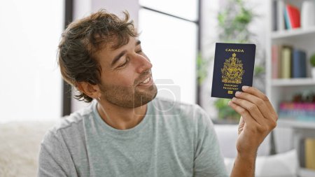 Téléchargez les photos : Heureux jeune homme, souriant et assis à la maison sur le canapé, tenant son passeport canadien, prêt pour les vacances! - en image libre de droit