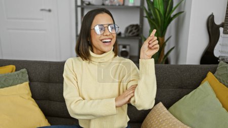 Téléchargez les photos : Joyeux jeune femme portant des lunettes assis sur un canapé dans un salon confortable intérieur, pointant et souriant. - en image libre de droit