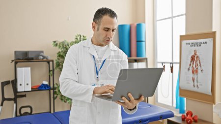 Photo for Hispanic male doctor using a laptop in a bright rehabilitation clinic room with physiotherapy equipment. - Royalty Free Image