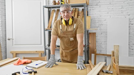 Téléchargez les photos : Un homme d'âge moyen dans un tablier se tient avec confiance dans un atelier de menuiserie lumineux entouré de bois, d'outils et d'équipements de sécurité. - en image libre de droit