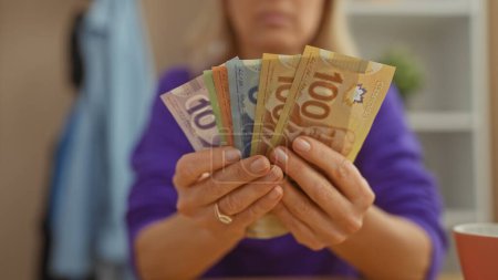Téléchargez les photos : Une femme dans un pull violet tenant la monnaie canadienne dans un cadre confortable à la maison avec un fond flou et dessus de table au premier plan. - en image libre de droit