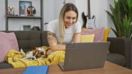 A smiling young woman works on her laptop at home with her small dog resting beside her on the couch.