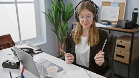 Téléchargez les photos : Une jeune femme professionnelle avec écouteurs dans un cadre de bureau moderne se concentrant sur une tâche à son bureau. - en image libre de droit
