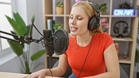 Photo for A blonde woman in a red shirt speaking into a microphone with headphones in a radio studio, clearly engaging in broadcasting or podcasting. - Royalty Free Image