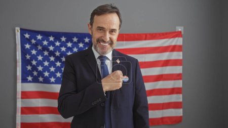 Smiling bearded man in suit displaying i voted sticker with american flag background in indoor setting.