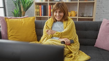 Foto de Mujer de mediana edad sonriente envuelta en una acogedora manta viendo la televisión en una elegante sala de estar. - Imagen libre de derechos