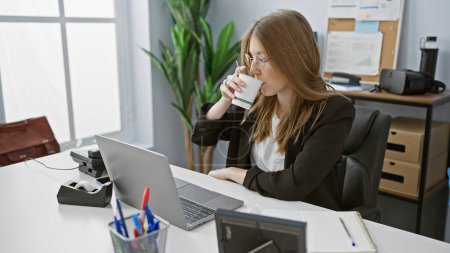 Eine junge erwachsene kaukasische Frau trinkt Kaffee in ihrem Büro mit moderner Ausstattung und Technik.