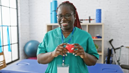 Photo for African american woman with braids smiling in scrubs holding a heart inside a bright physiotherapy clinic. - Royalty Free Image
