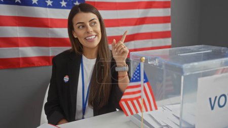Foto de Mujer joven estadounidense señalando en el interior de la universidad electoral con bandera, boleta electoral, etiqueta de voto y expresión alegre. - Imagen libre de derechos
