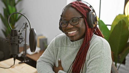 Téléchargez les photos : Femme afro-américaine avec des tresses et des lunettes souriant dans un studio de radio portant un casque - en image libre de droit