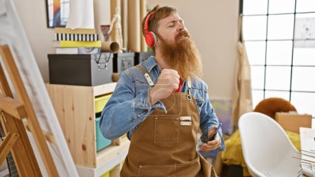 Handsome, bearded, young redhead man who is an artist, dancing while listening to music in art studio, canvas, paintbrush in hand, smiling with joy.