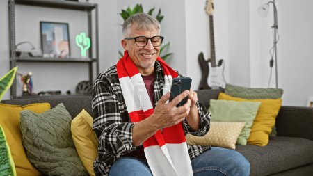 Photo for A cheerful middle-aged man with grey hair using a smartphone at home, seated on a grey couch surrounded by colorful pillows. - Royalty Free Image