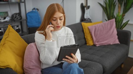 Foto de Una pelirroja enfocada en casa multitarea con una llamada telefónica y una tableta en una acogedora sala de estar. - Imagen libre de derechos