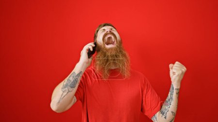 Téléchargez les photos : Jeune homme rousse ravi de célébrer extatiquement tout en parlant au téléphone sur un fond rouge isolé - en image libre de droit