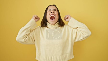Téléchargez les photos : Jeune femme hispanique énergique acclamant dans un pull crème sur un fond jaune vif. - en image libre de droit