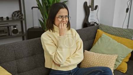 Téléchargez les photos : Une femme hispanique coûteuse portant des lunettes et un pull jaune s'assoit délicatement sur un canapé gris à l'intérieur. - en image libre de droit