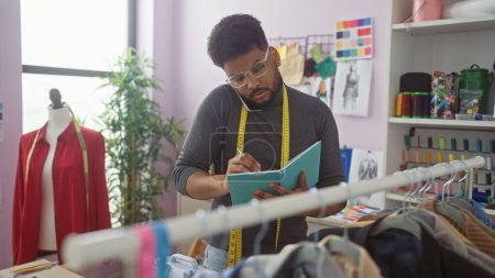 Hombre afroamericano multitarea en una sastrería con teléfono y bloc de notas, rodeado de prendas y artículos de costura.