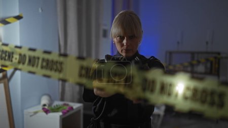 A serious middle-aged female police officer examines a crime scene with caution tape in an indoor setting.