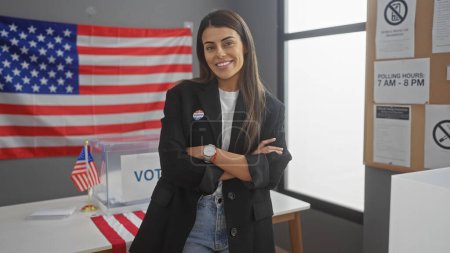 Foto de Una mujer hispana confiada se levanta con los brazos cruzados en una sala del colegio electoral americano con una bandera y signos de voto. - Imagen libre de derechos