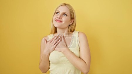 Grateful young woman with blonde hair and a heartfelt expression standing against a yellow backdrop