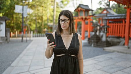 Foto de Hermosa mujer hispana en gafas, totalmente absorta en su teléfono, atrapada en medio de escribir un texto emocionante en fushimi inari taisha, un icónico santuario kyoto, inmerso en la cultura japonesa. - Imagen libre de derechos