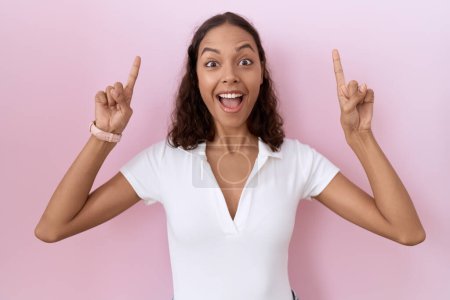 Foto de Young hispanic woman wearing casual white t shirt smiling amazed and surprised and pointing up with fingers and raised arms. - Imagen libre de derechos