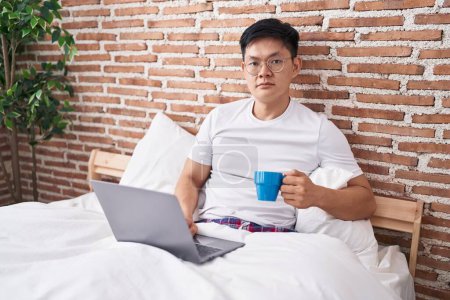 Photo for Young asian man drinking coffee sitting on the bed thinking attitude and sober expression looking self confident - Royalty Free Image