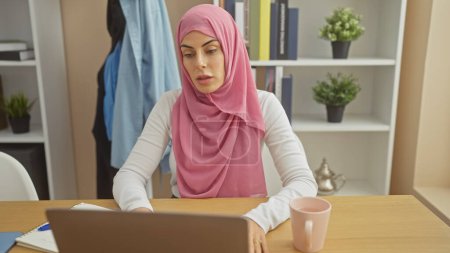 Téléchargez les photos : Une femme concentrée portant un hijab rose travaille sur son ordinateur portable à un bureau en bois, avec des plantes et une théière dans son bureau à la maison soigné. - en image libre de droit