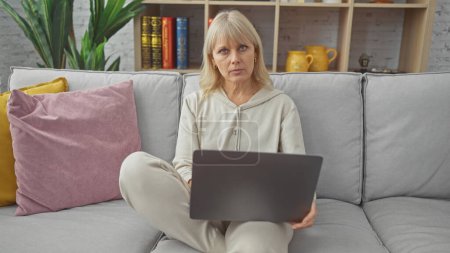 Téléchargez les photos : Une femme blonde utilise un ordinateur portable assis sur un canapé dans un salon confortable, dépeignant le travail à distance ou les loisirs. - en image libre de droit