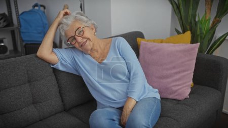 Photo for A smiling elderly woman with grey hair wearing glasses relaxes on a couch in a cozy living room. - Royalty Free Image
